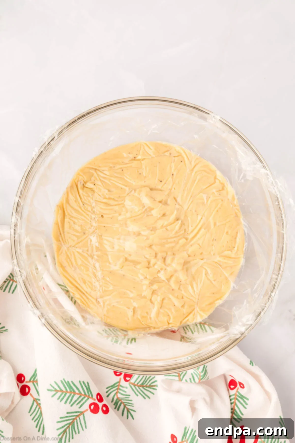 A medium bowl filled with warm eggnog pudding, covered tightly with plastic wrap pressed directly onto its surface to prevent a film from forming during refrigeration.