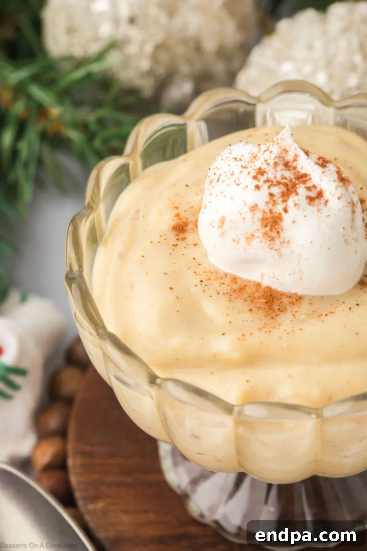 A close-up of a glass bowl holding rich eggnog pudding, lavishly topped with a swirl of fresh whipped cream and a delicate dusting of ground cinnamon, ready for a festive occasion.