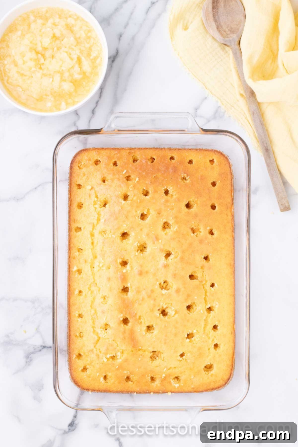 A baked yellow cake in a baking dish, its surface dotted with numerous holes, ready to absorb the juicy pineapple and creamy pudding layers.