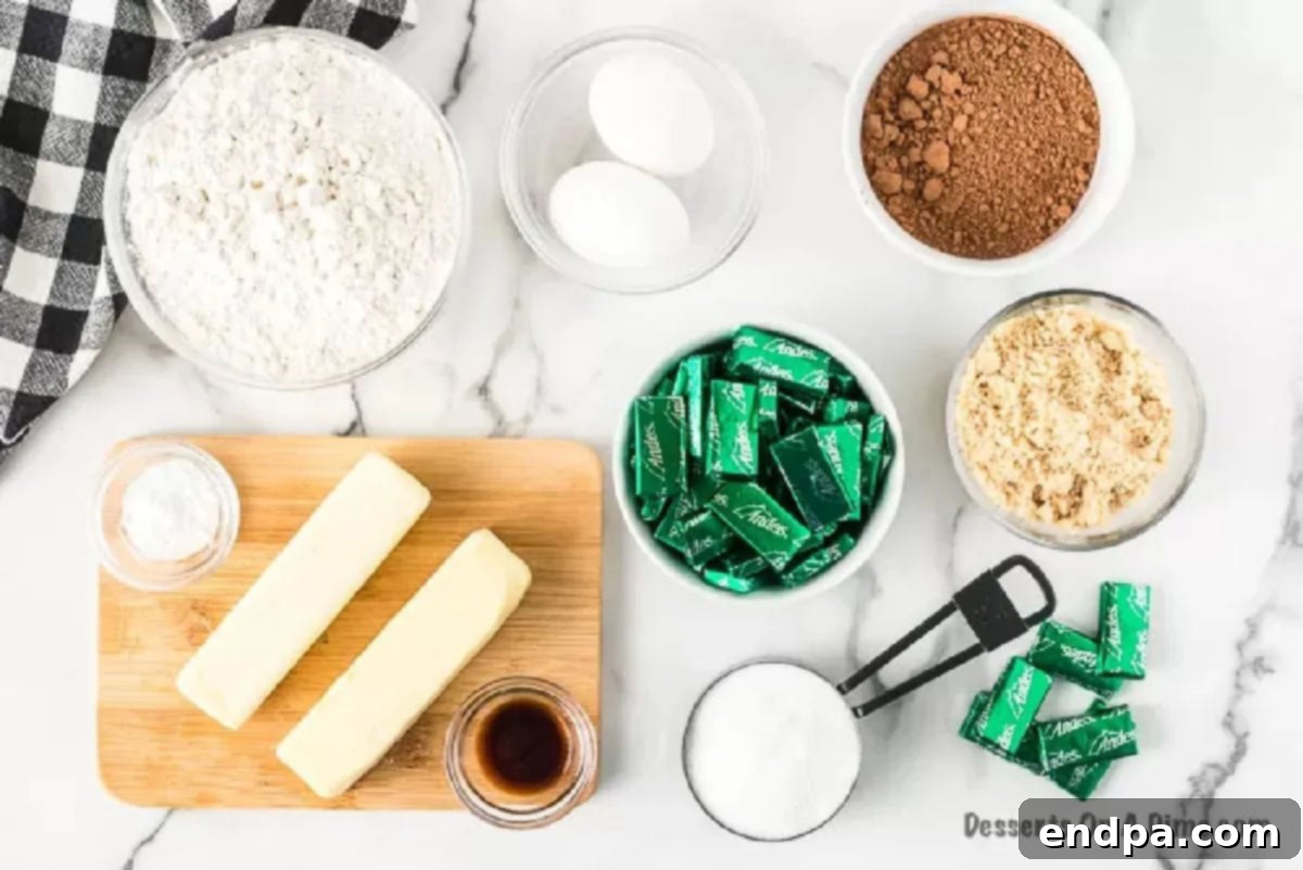 Key ingredients for Andes Mint Cookies laid out: softened butter, granulated sugar, brown sugar, fresh eggs, vanilla extract, all-purpose flour, cocoa powder, baking soda, salt, and a pile of Andes Mint candies.