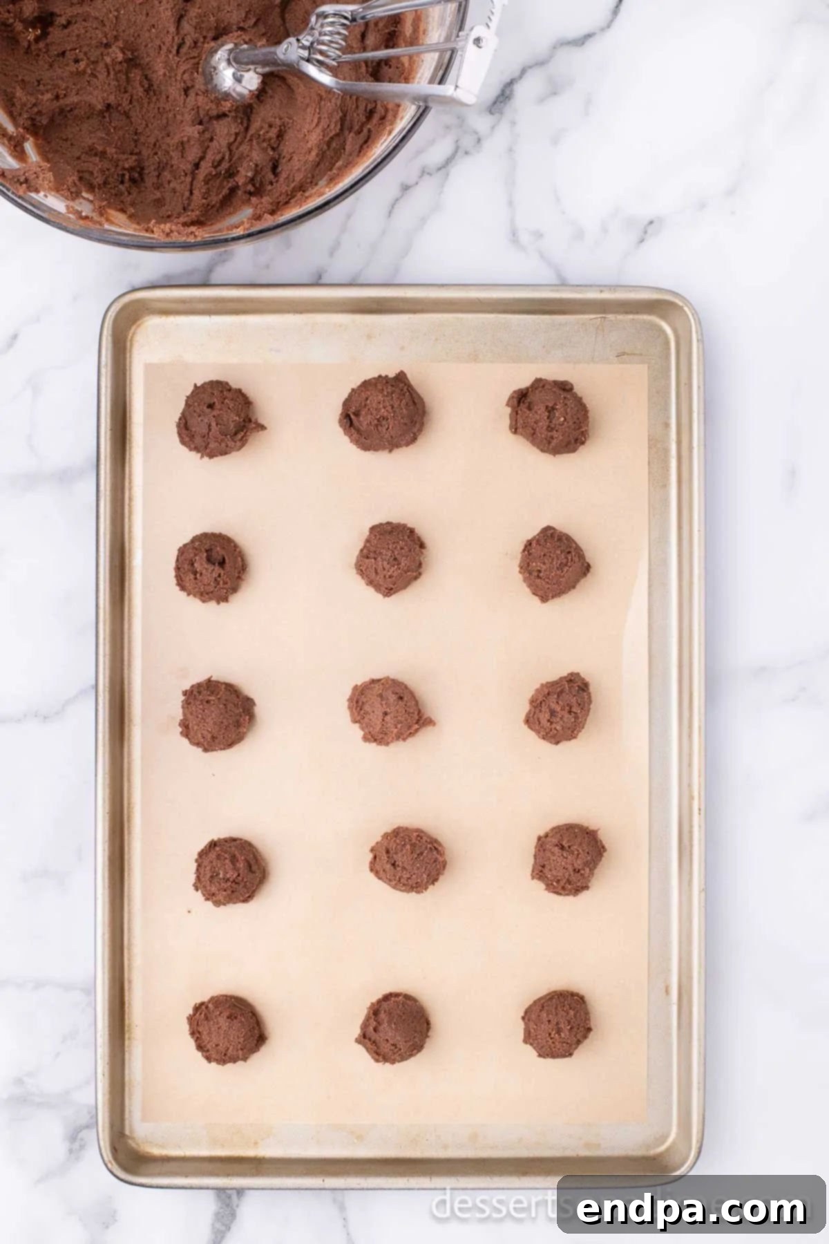 Uniform balls of chocolate cookie dough are neatly scooped onto a baking sheet lined with a silicone mat, spaced adequately for baking.