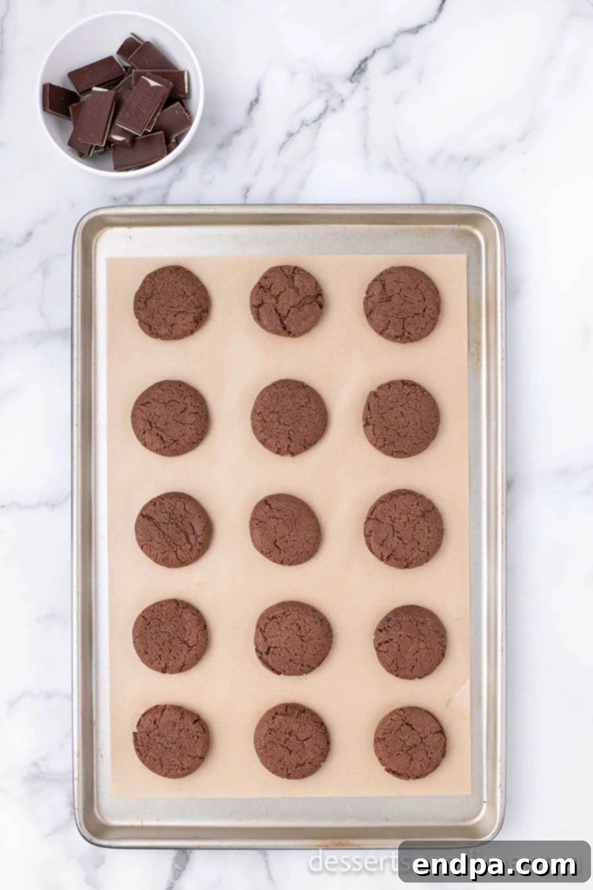 Freshly baked chocolate cookies are cooling on a baking sheet, with their edges set but centers still slightly soft.