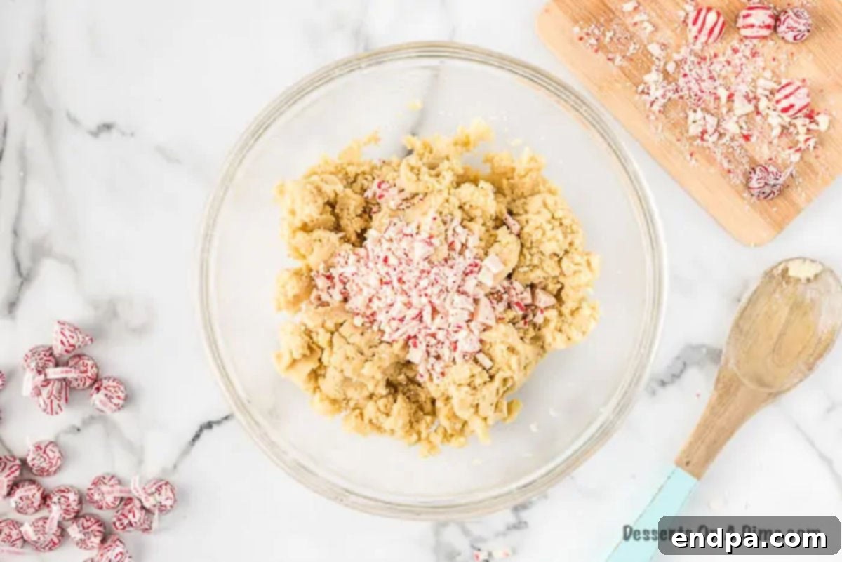 A mixing bowl showing combined cookie batter with visible pieces of chopped Hershey's Candy Cane Kisses folded in, ready for rolling.