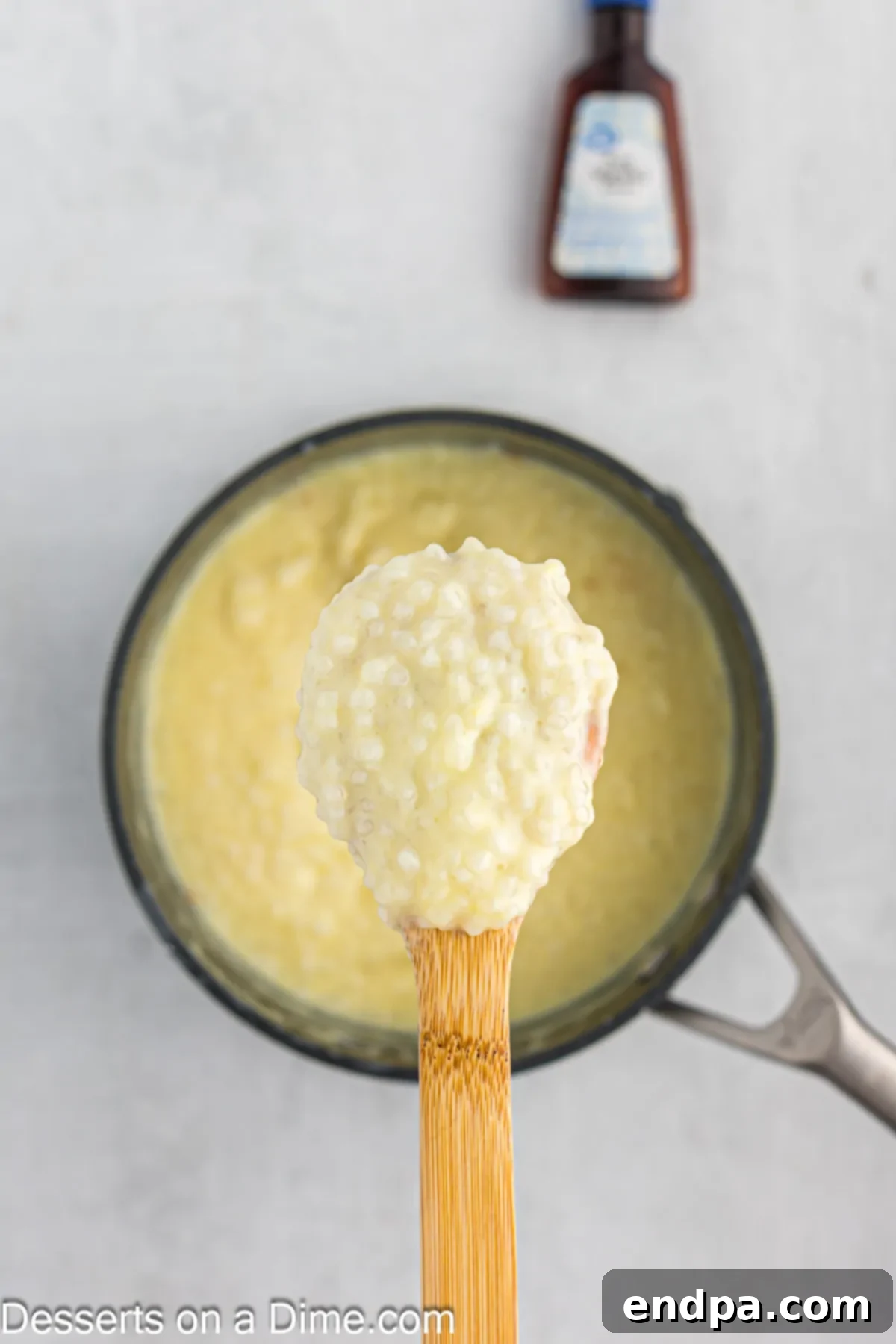 A wooden spoon stirring the thickened tapioca pudding in the saucepan, with vanilla extract just added for aroma.