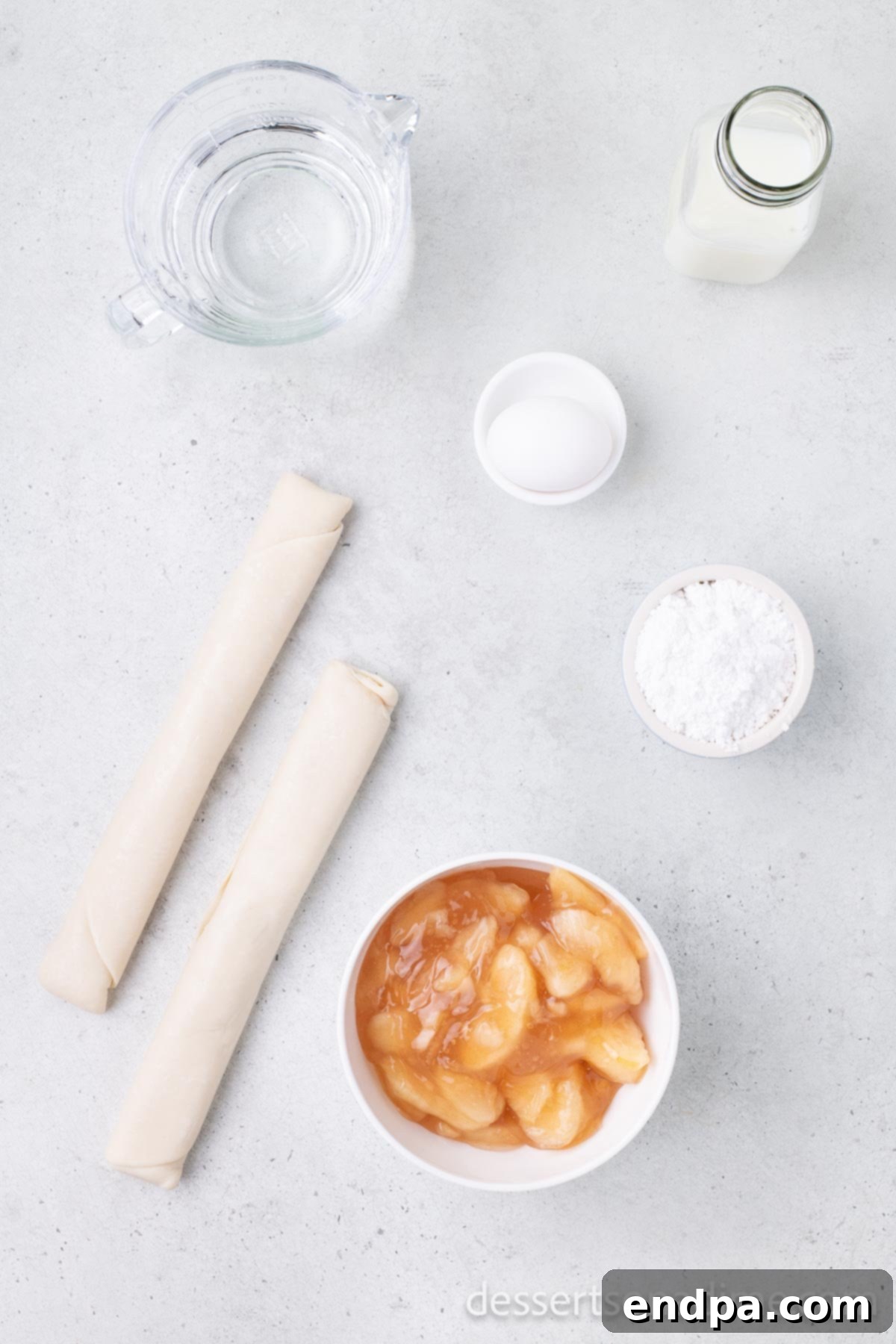 Arrangement of ingredients for air fryer hand pies: refrigerated pie crust, apple pie filling, a large egg, powdered sugar, and a small bowl of milk.