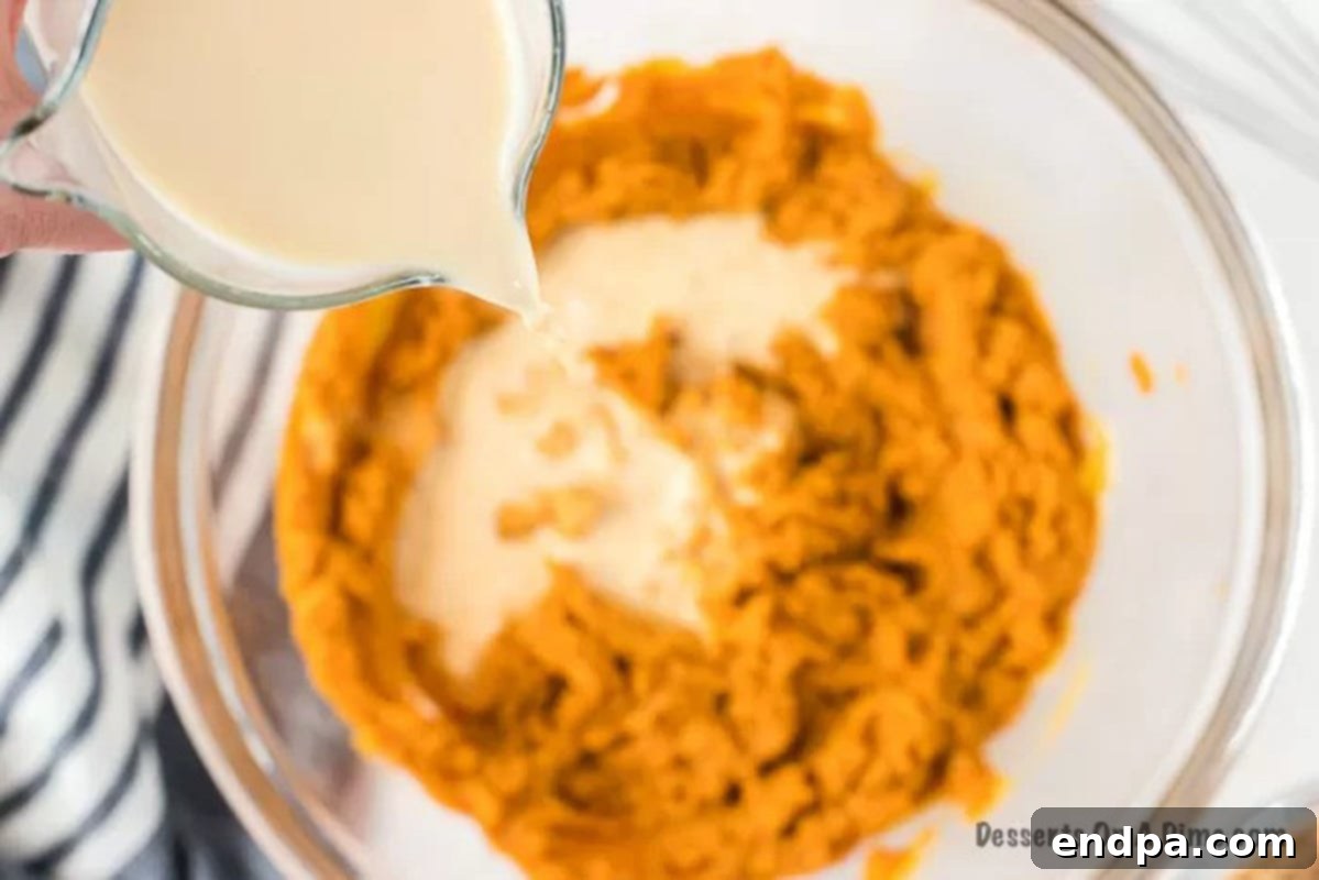 Evaporated milk is slowly poured into the pumpkin pie mixture in a large bowl, being gently stirred to achieve a smooth, creamy consistency.