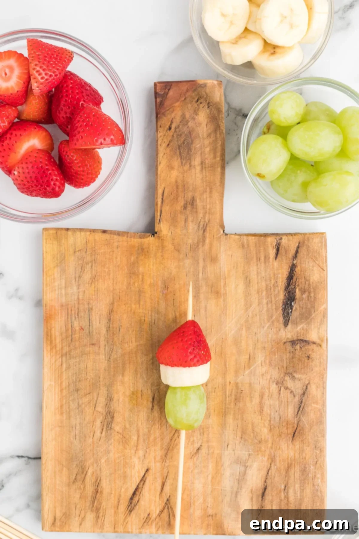 A prepared strawberry threaded onto the skewer above the banana, completing the body of the Grinch's Santa hat.