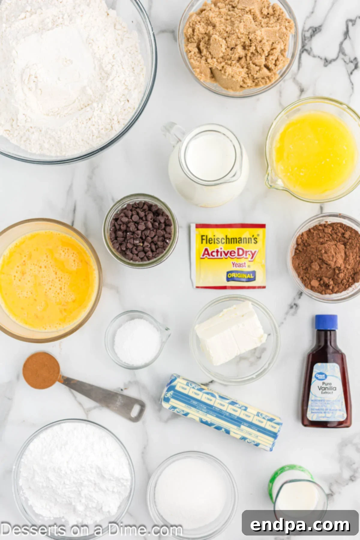 Ingredients for Chocolate Chip Cinnamon Rolls laid out on a table: butter, yeast, flour, sugar, chocolate chips, vanilla, cocoa powder.