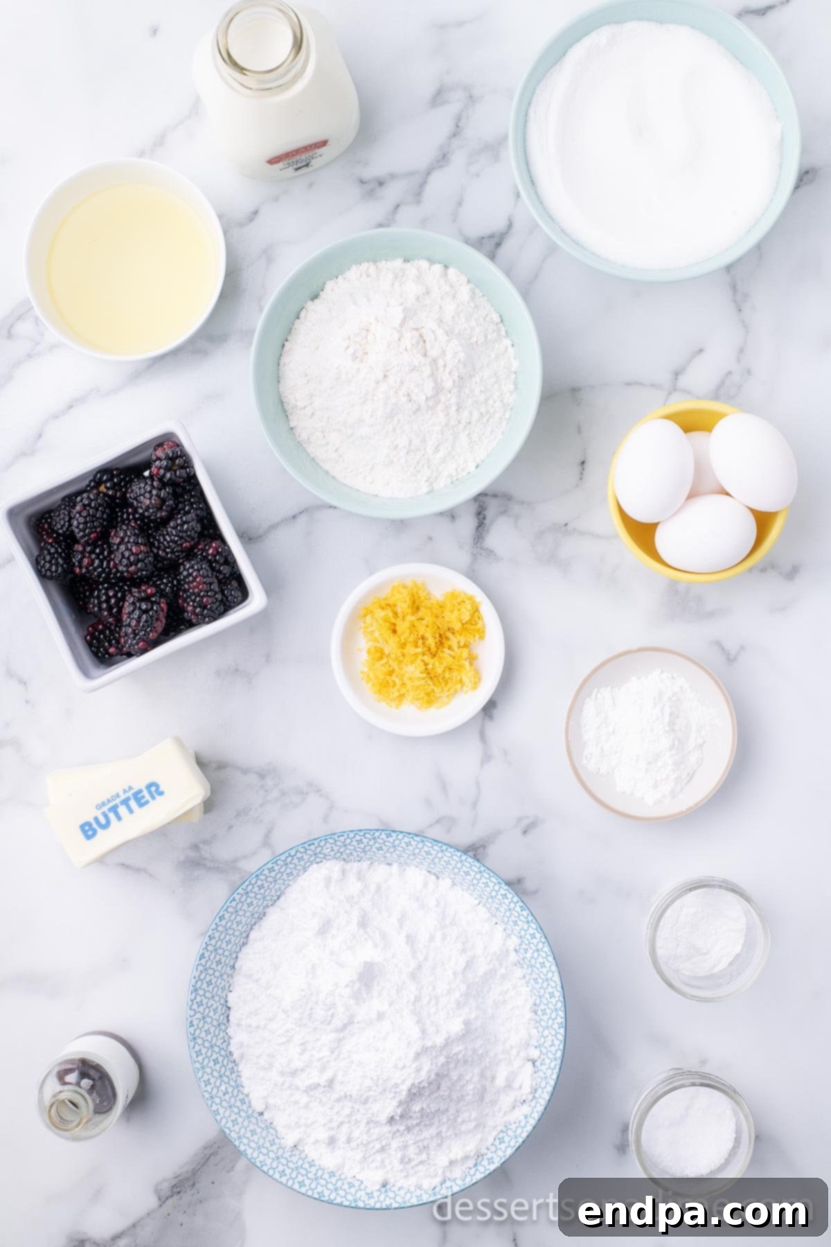 Luscious Lemon Blackberry Cake 3 A flat lay photograph of various ingredients laid out for baking a lemon blackberry cake, including flour, baking powder, eggs, whole milk, fresh lemons, vanilla extract, unsalted butter, granulated sugar, fresh blackberries, powdered sugar, and cornstarch.