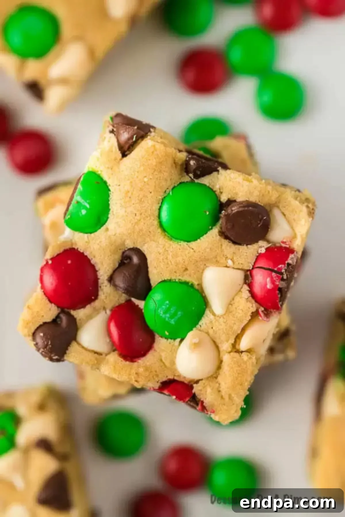 Christmas cookie bars stacked, loaded with chocolate chips, M&M's, and a gooey texture for a decadent holiday treat. Another view of stacked chocolate chip cookie bars, emphasizing their chewy texture and festive appearance.