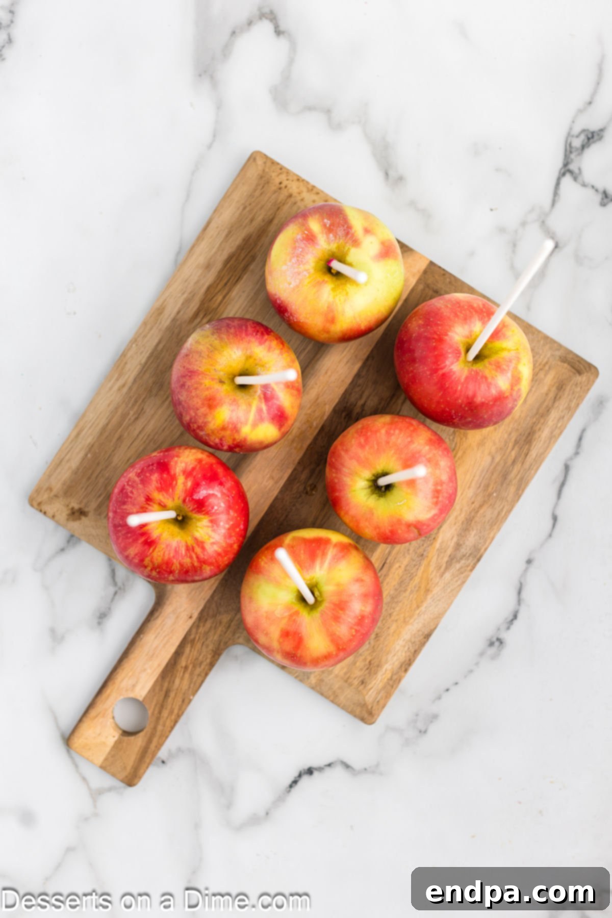 Apples on a cutting board with wooden sticks securely inserted into their stem ends, ready for dipping.