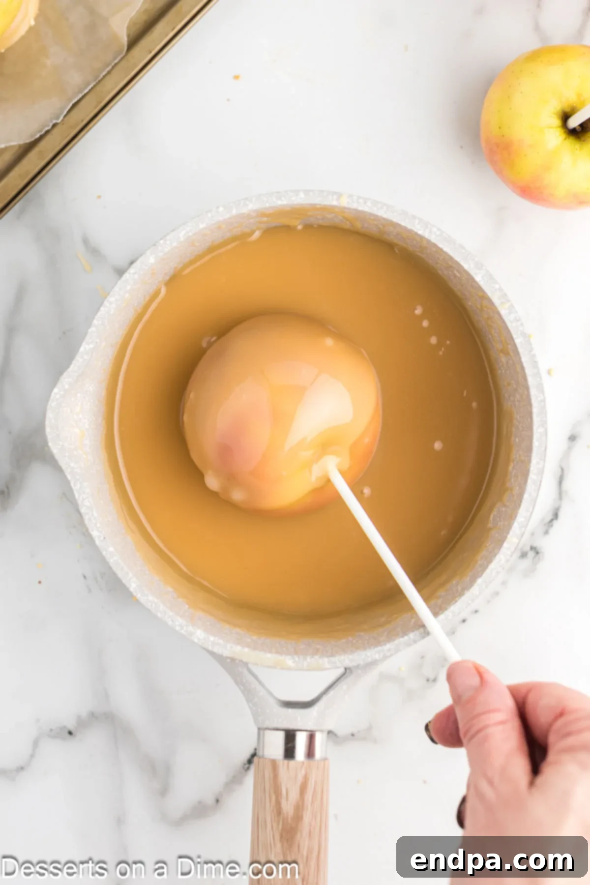 Excess caramel gracefully dripping off a freshly coated apple held over the saucepan, ensuring a smooth finish.