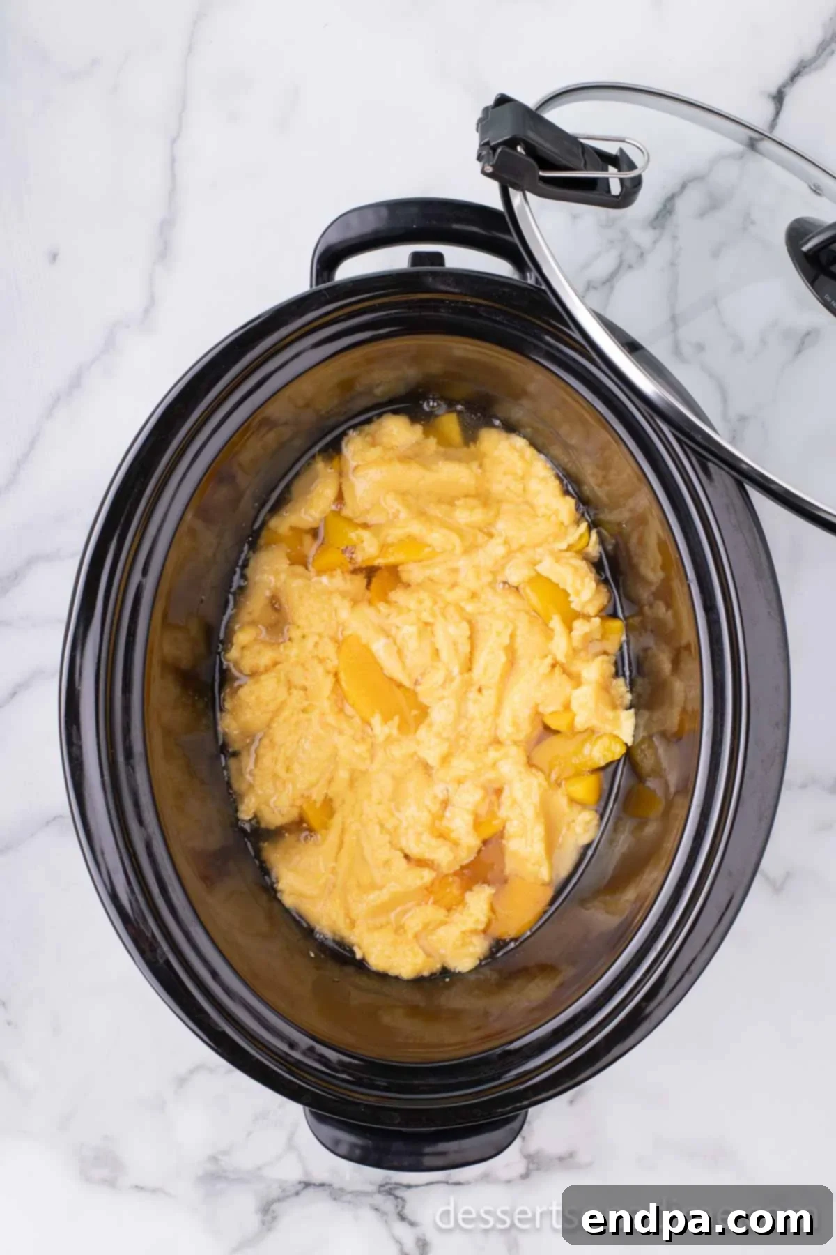 The butter-infused cake mix is being evenly spread over the layer of peaches in the slow cooker, forming the top layer of the dump cake.