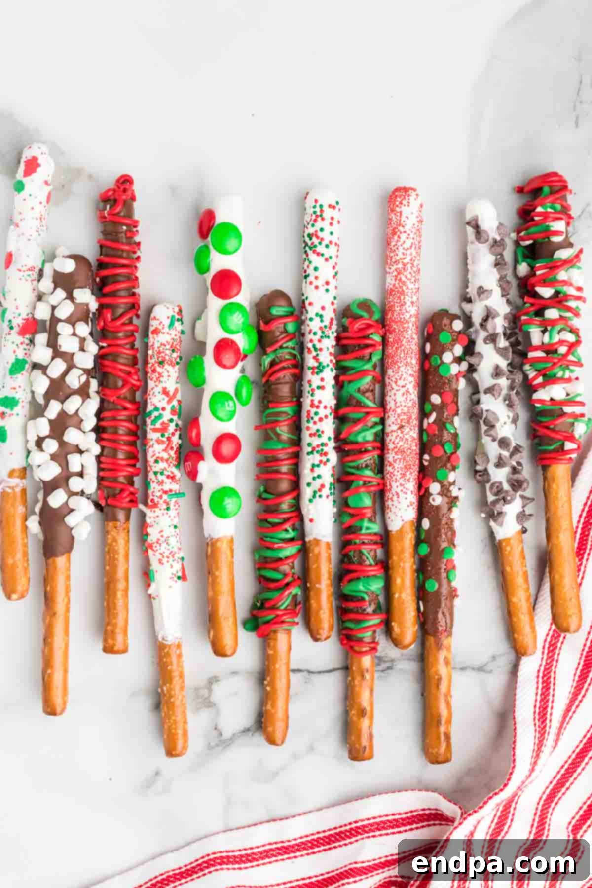 Crispy pretzel rods generously dipped in vibrant Christmas-themed chocolate and adorned with an assortment of festive sprinkles, colorful M&Ms, and crunchy chocolate chips, laid out on a baking sheet.