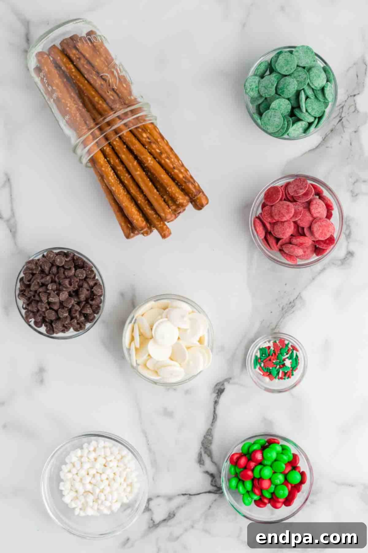 A flat lay photograph displaying the simple ingredients needed for Christmas Chocolate Covered Pretzel Rods: a bag of pretzel rods, colorful candy melts, a bowl of semi-sweet chocolate chips, and an assortment of festive sprinkles and M&Ms.