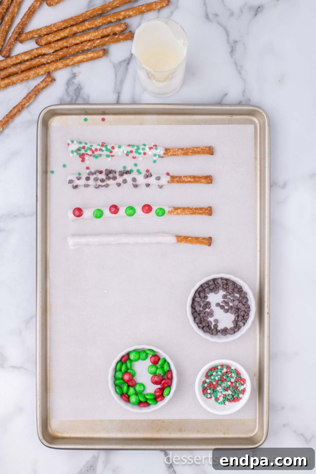 A close-up view of chocolate-dipped pretzel rods adorned with festive Christmas sprinkles, resting on a parchment-lined baking sheet.
