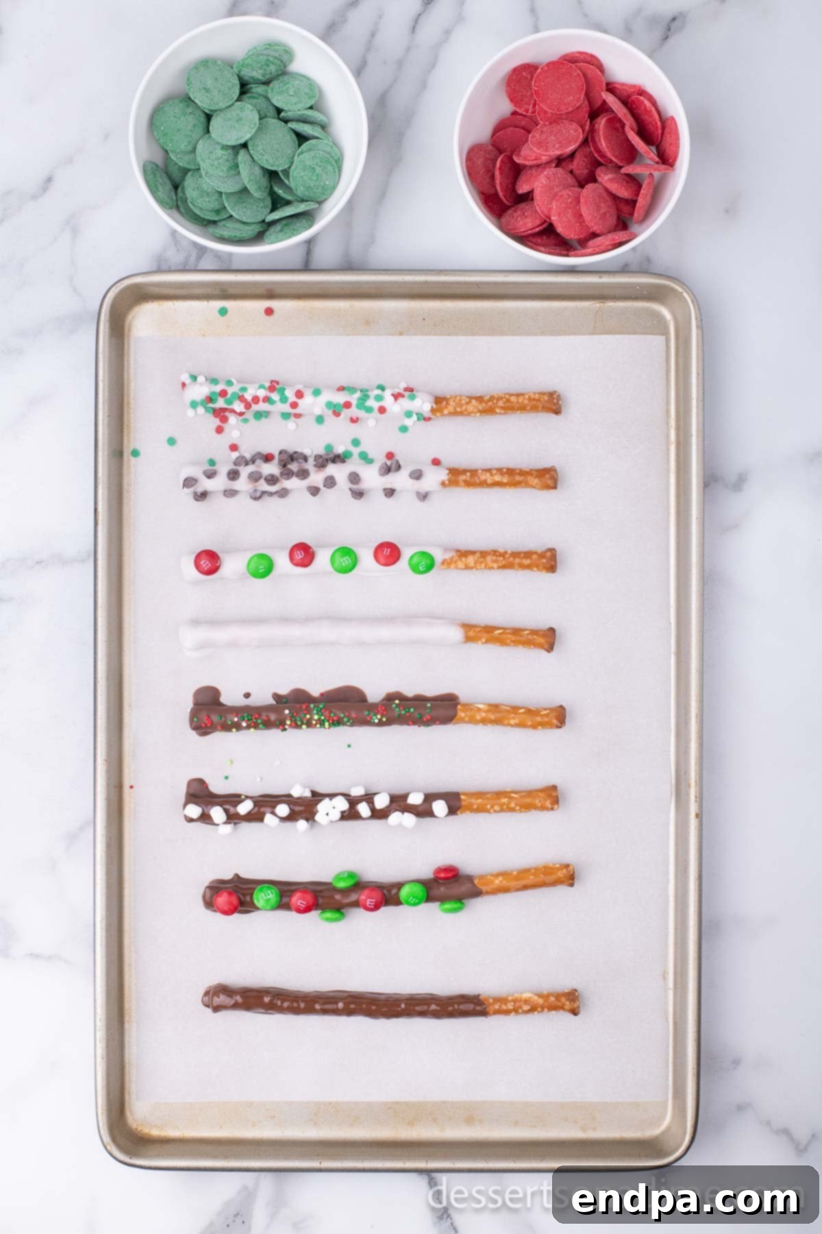 A vibrant assortment of chocolate-covered pretzel rods on a baking sheet, showcasing various Christmas-themed toppings including sprinkles, M&Ms, and crushed candy canes.