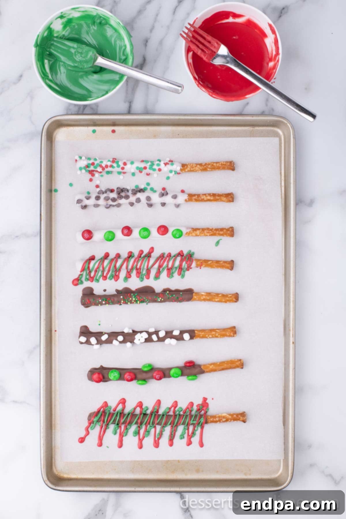 An array of elaborately decorated Christmas pretzel rods, featuring intricate drizzles of colored candy melts and a generous coating of festive sprinkles, arranged on a baking sheet.