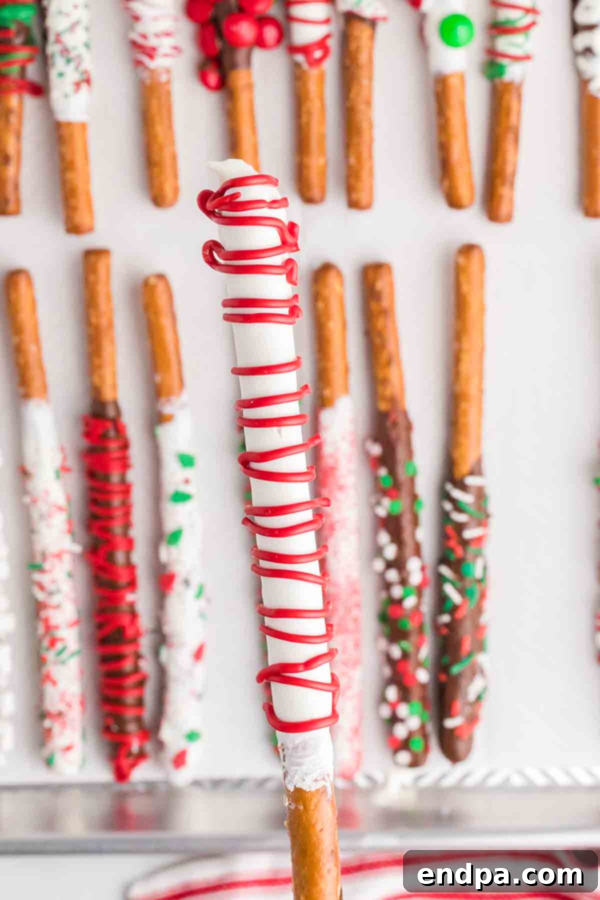 A beautifully decorated Christmas Chocolate Covered Pretzel Rod, featuring a red candy melt base with delicate white drizzles and sparkling festive sprinkles, presented upright.