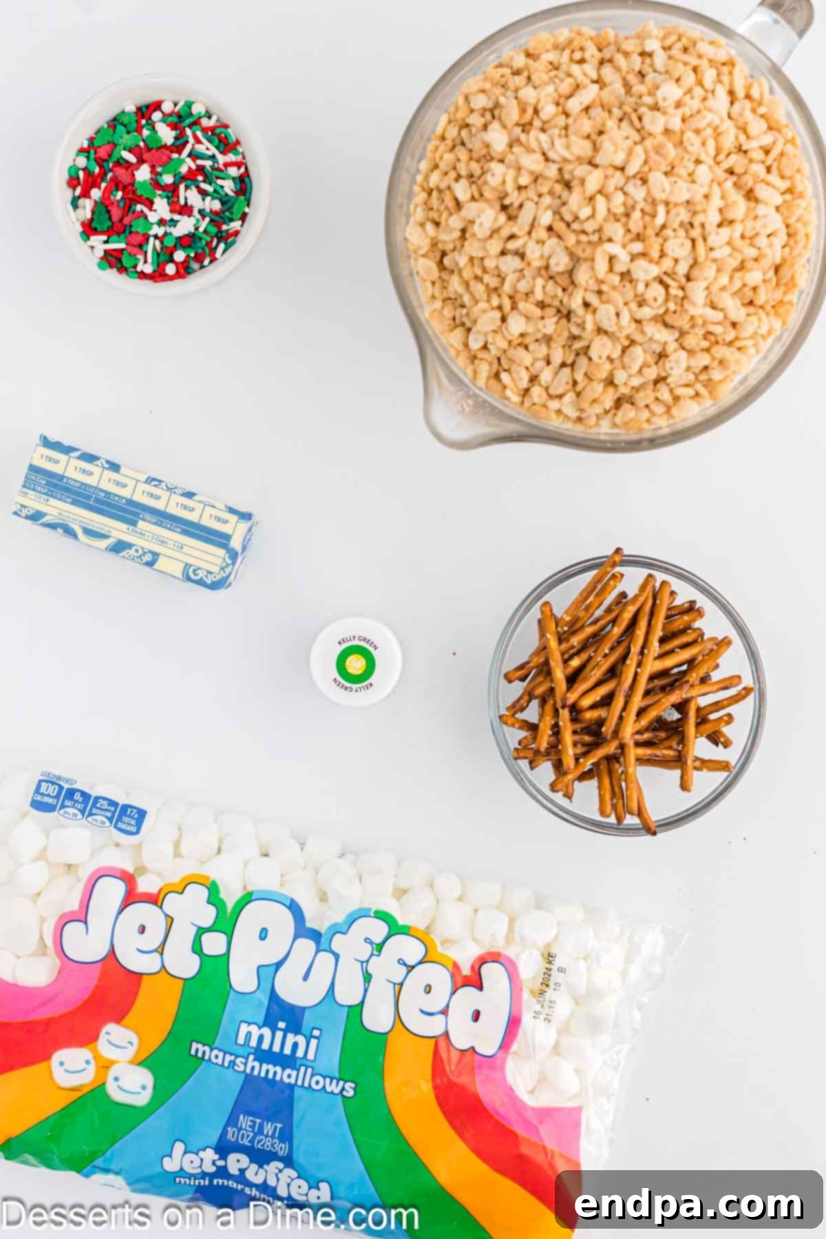 A collection of key ingredients for Christmas Tree Rice Krispie Treats, including butter, mini marshmallows, green food coloring, festive sprinkles, and pretzel sticks.