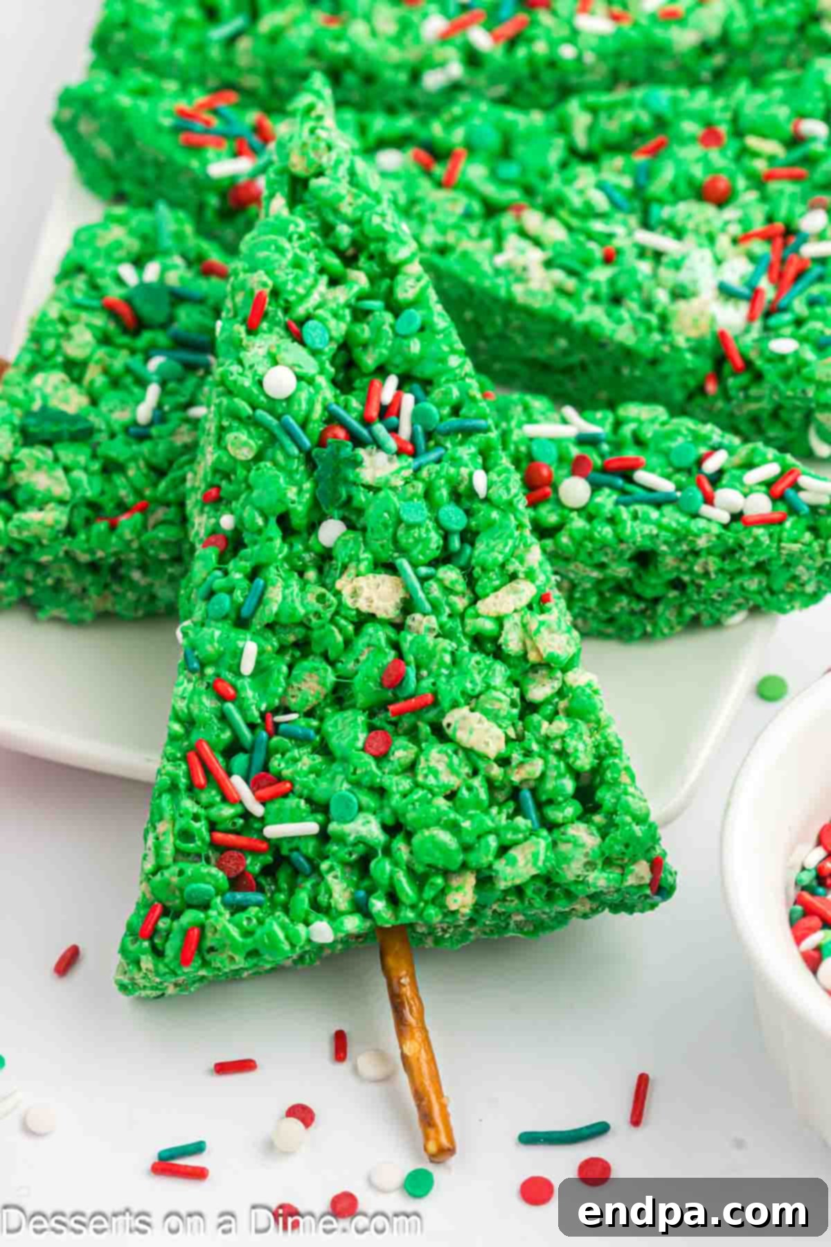 Triangle shapes cut from the Rice Krispie mixture, each with a pretzel stick inserted at the bottom to form a tree trunk.