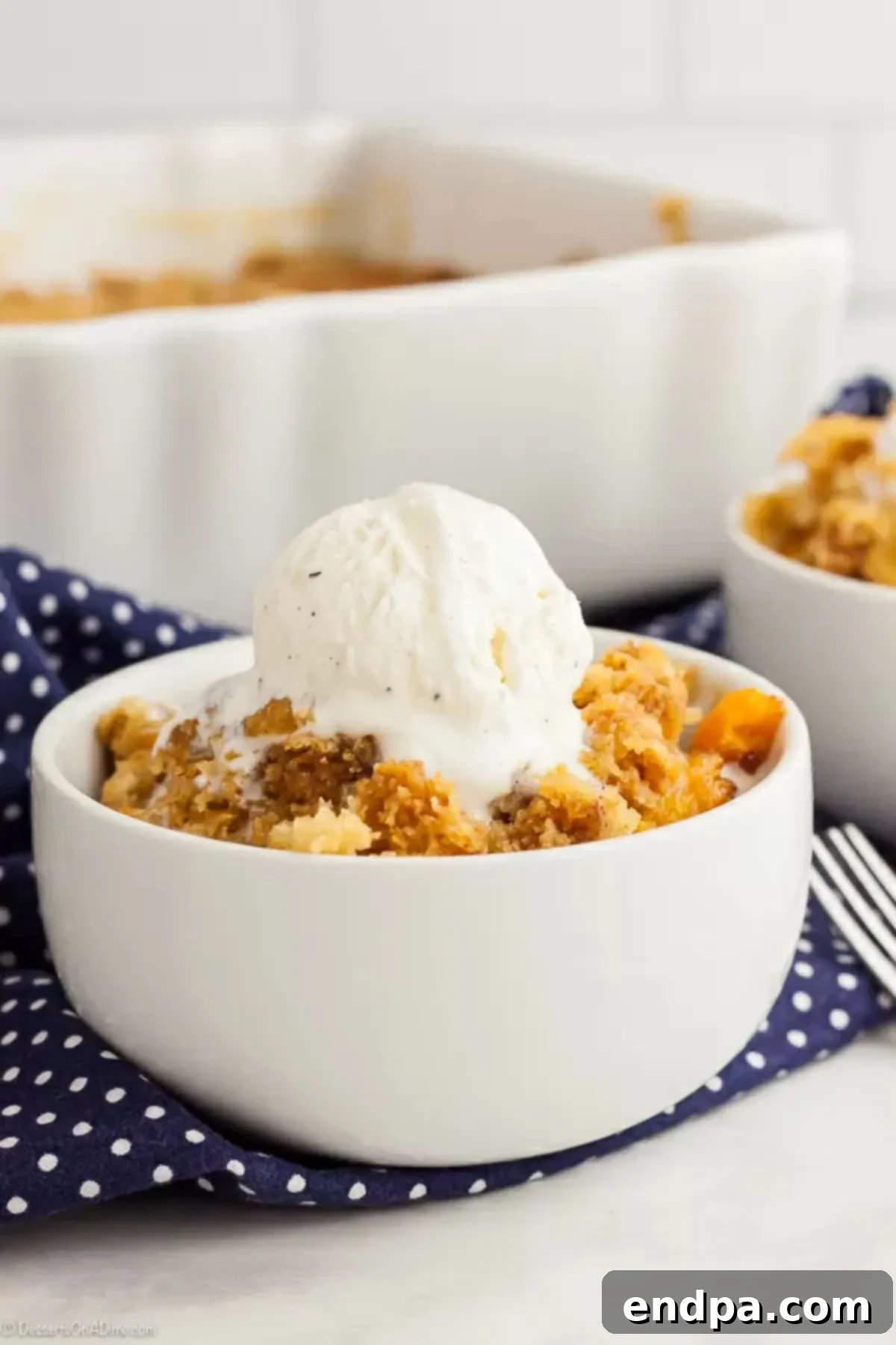 White bowl with peach dump cake topped with a scoop of vanilla ice cream, ready to be enjoyed.