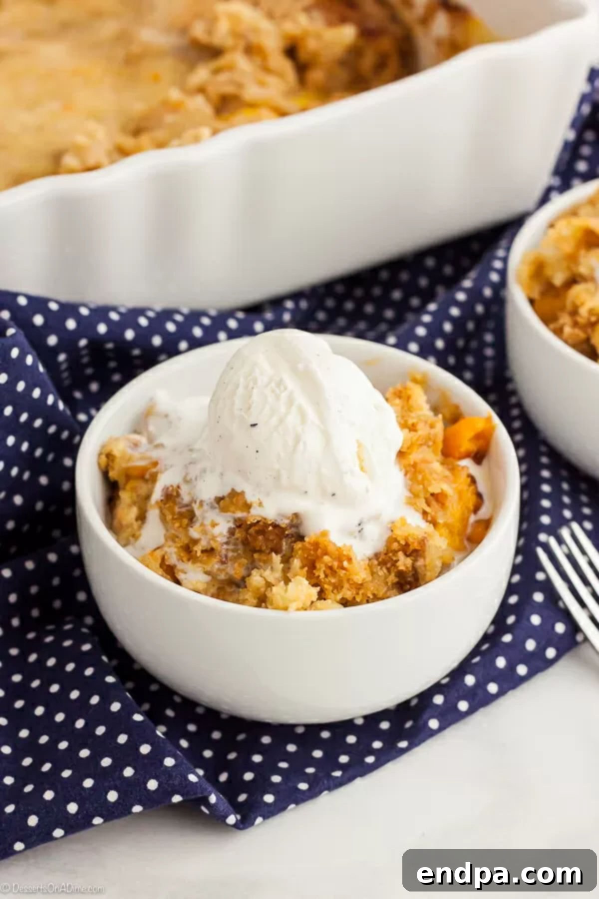 A close-up of a white bowl with peach dump cake, generously topped with melting vanilla ice cream and a spoon.