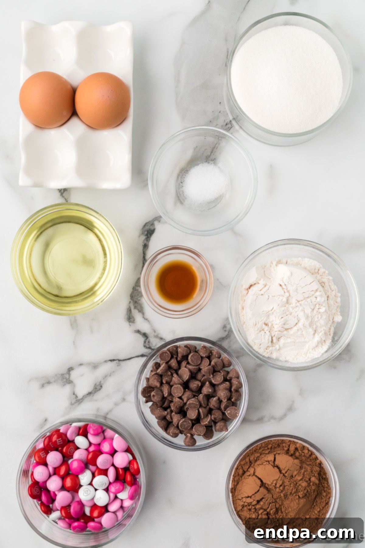 Ingredients laid out for homemade brownies: granulated sugar, vegetable oil, vanilla extract, cocoa powder, all-purpose flour, chocolate chips, and festive M&Ms.