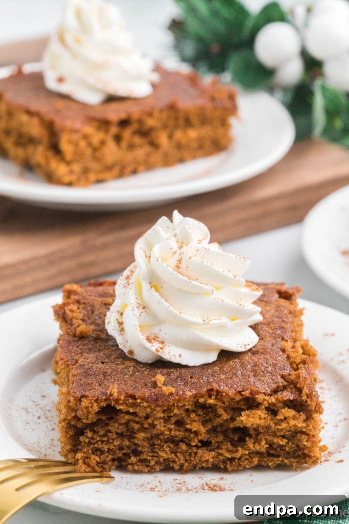 A perfectly sliced piece of gingerbread cake on a white plate, elegantly topped with a generous dollop of fluffy whipped cream and a sprinkle of powdered sugar, ready to be enjoyed.