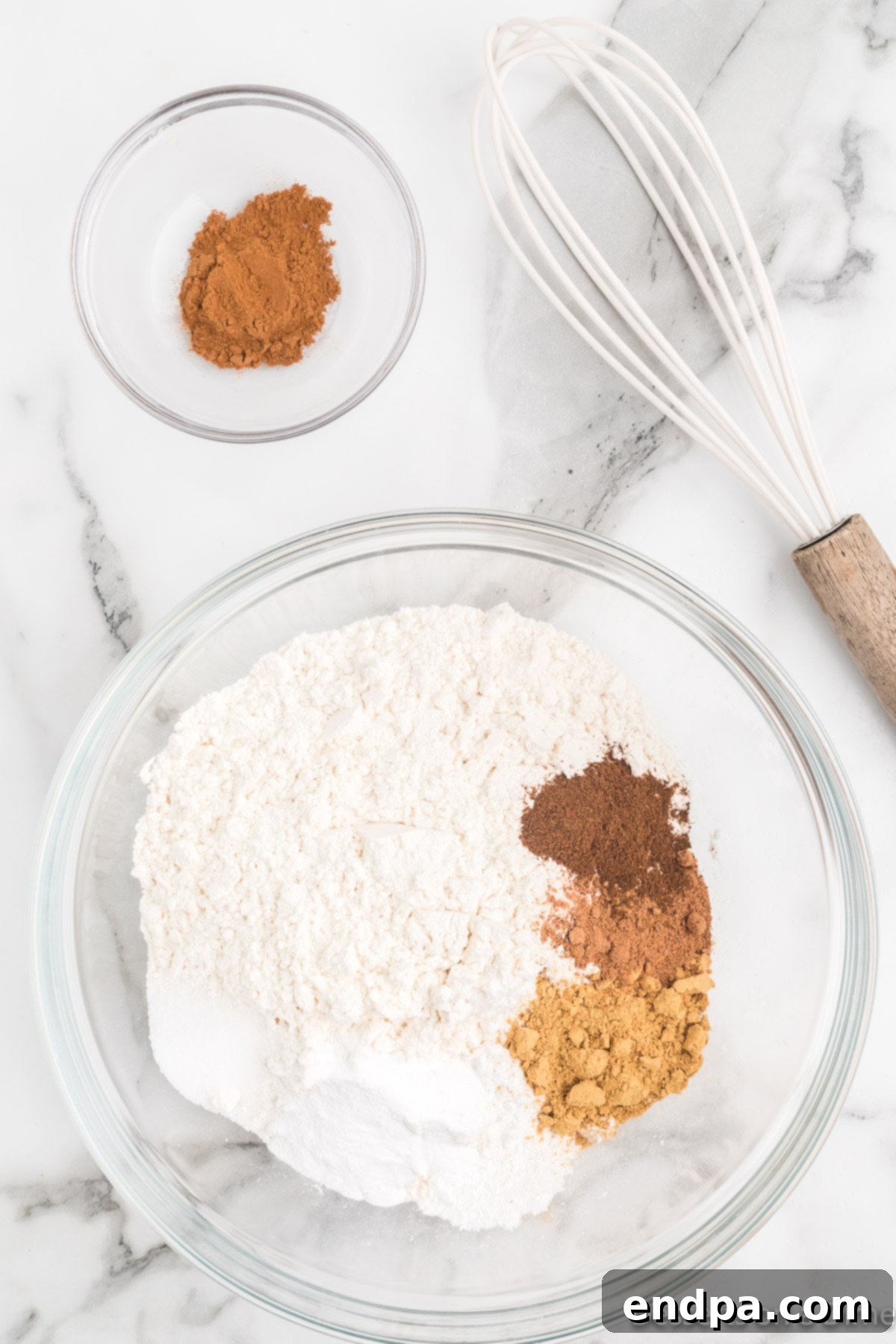 A medium mixing bowl containing white flour, baking powder, baking soda, salt, ground ginger, ground cinnamon, ground nutmeg, and ground cloves, with a whisk resting in the bowl, ready to combine the dry ingredients for the gingerbread cake.