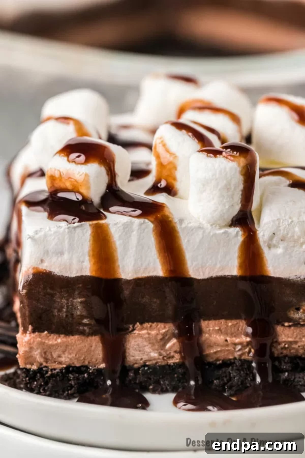Decadent Hot Cocoa Lasagna 2 Close up of a piece of hot chocolate lasagna on a small white plate with chocolate syrup drizzled on top of it. The background shows the full lasagna in a baking dish.