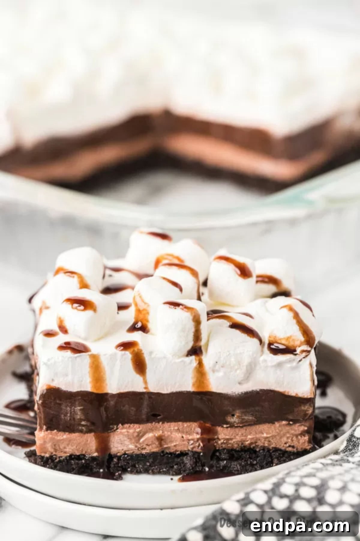 From the oreo crust to the decadent chocolate cheesecake mousse, this hot chocolate lasagna does not disappoint. The entire recipe for Hot chocolate lasagna dessert is so easy! Try this no bake dessert recipe. #dessertonadime #hotchocolatelasagna #christmasdesserts #cake #dessert #recipe #hotchocolatelasagnarecipe A piece of hot chocolate lasagna on a small white plate with the remaining lasagna in a pan behind it, showcasing its delicious appeal.