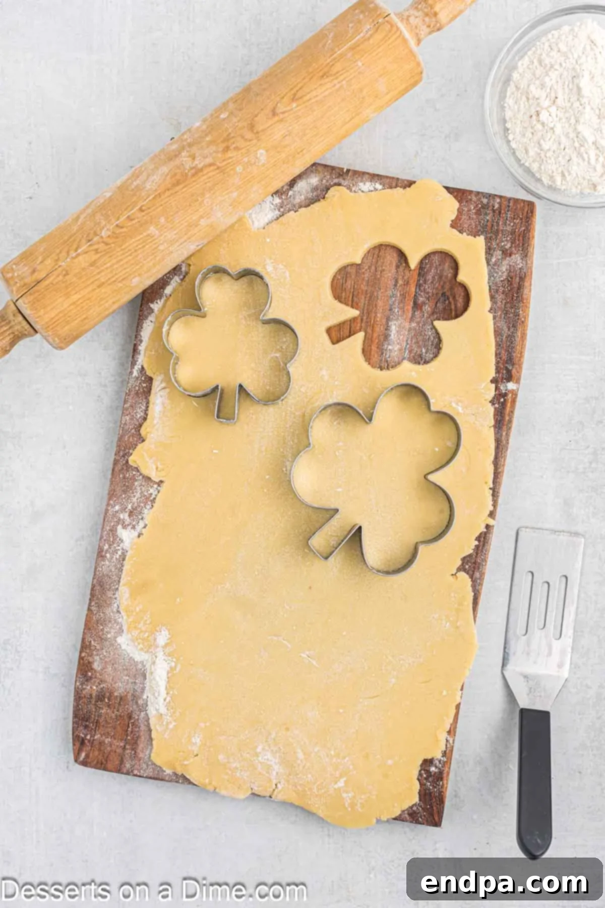 Emerald Clover Delights 8 Shamrock-shaped cookies neatly cut out from rolled dough on a lightly floured cutting board, ready for baking.