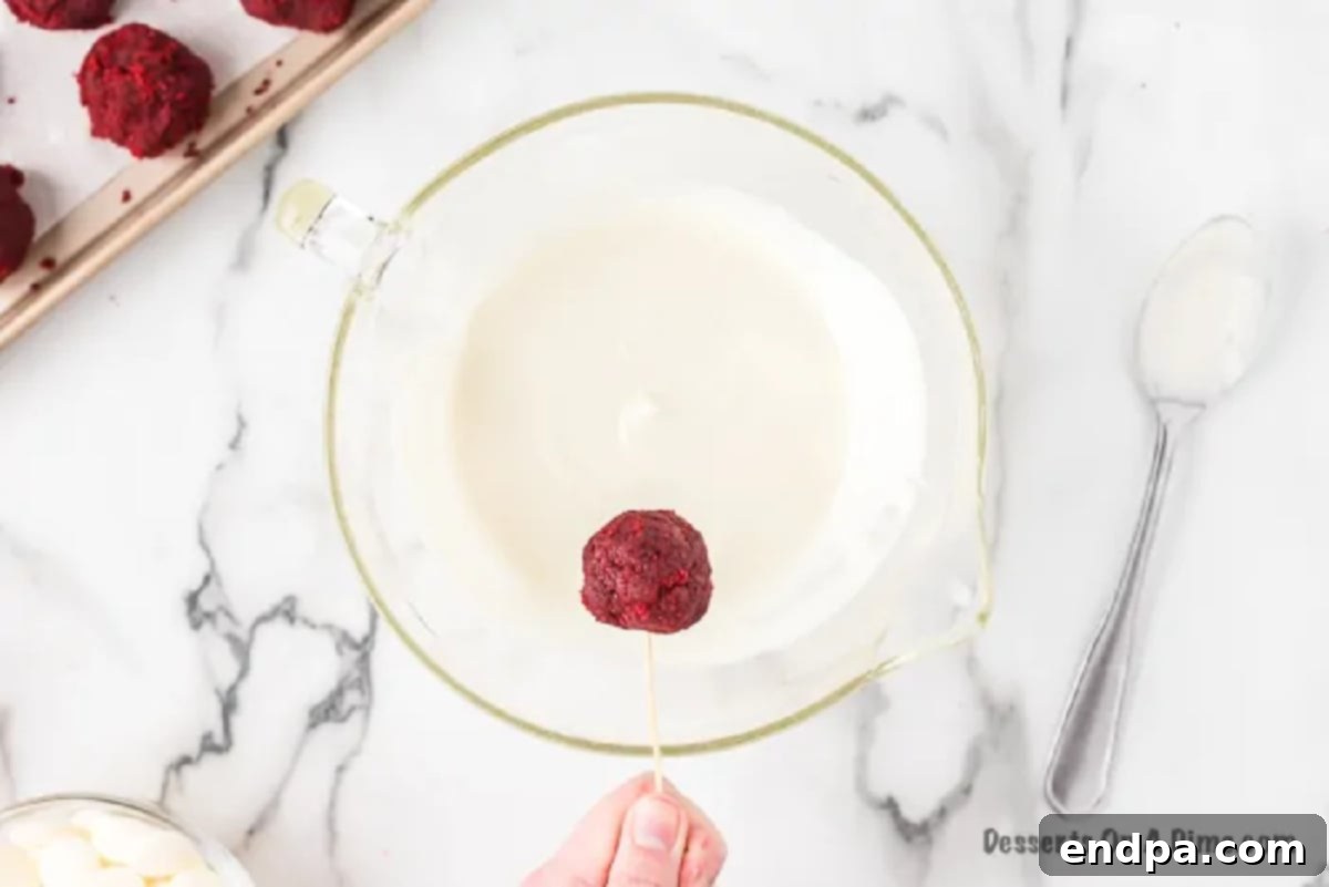 A red velvet cake ball being dipped into melted white chocolate with a toothpick.