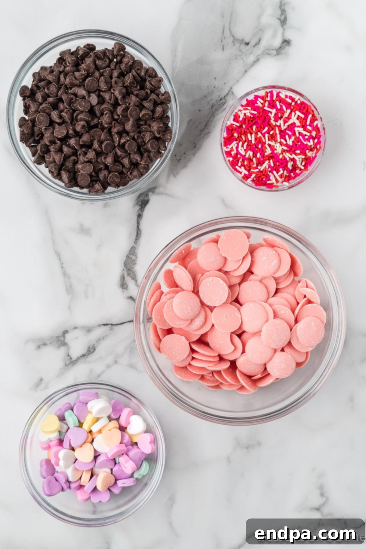 A collection of Valentine's Day bark ingredients, including chocolate chips, pink candy melts, conversation hearts, and sprinkles.