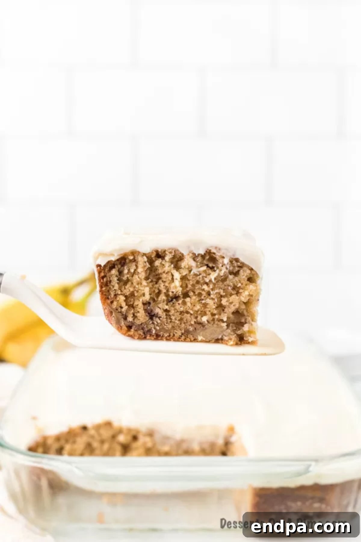 Foolproof Ultra-Moist Banana Cake 2 A beautifully frosted banana cake in a rectangular pan, with a perfect slice removed on a spatula, ready to be served.