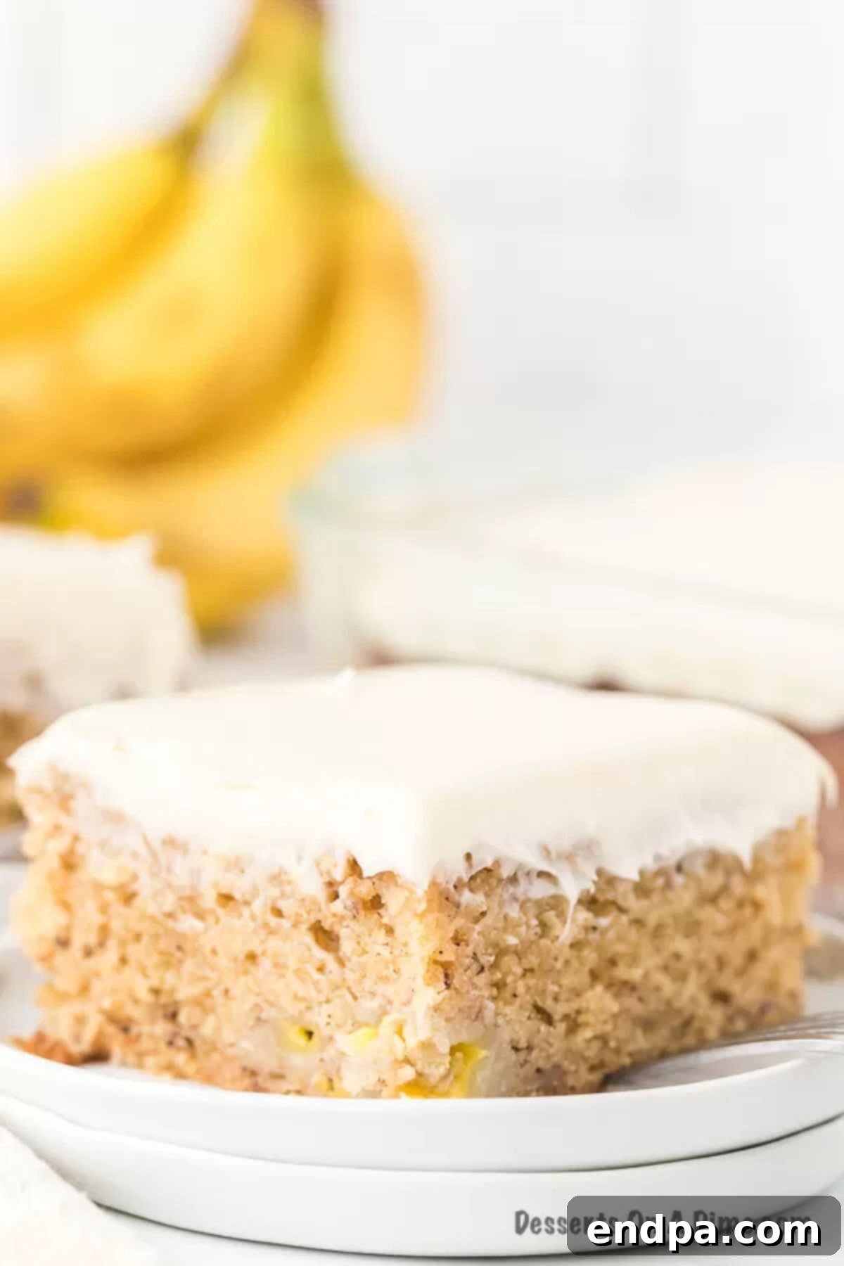 Foolproof Ultra-Moist Banana Cake 4 A delectable slice of banana cake topped with creamy white cream cheese frosting, presented on a simple white plate, ready to be enjoyed.