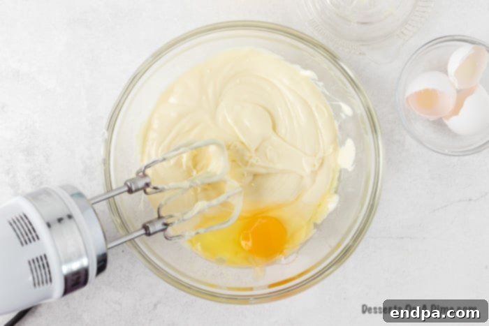 Zesty Lemon Cheesecake Bars 7 Eggs being added one at a time to the cheesecake filling in a bowl, with a mixer.