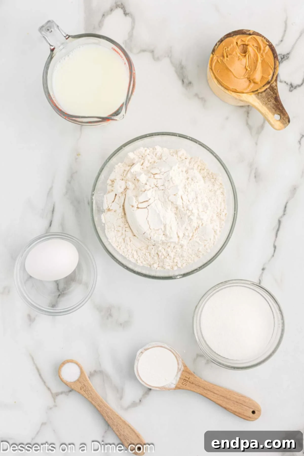 All the basic ingredients neatly laid out on a table: a bowl of flour, sugar, a single egg, baking powder, salt, a glass of milk, and a jar of creamy peanut butter.