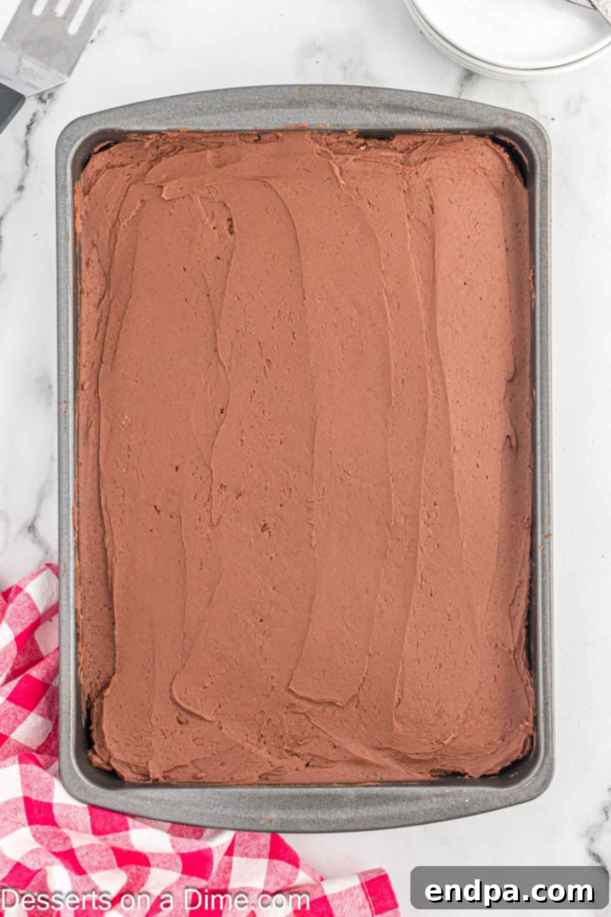 Effortless Decadent Chocolate Cake 9 Rich chocolate buttercream frosting being evenly spread across the cooled chocolate cake in a baking pan.