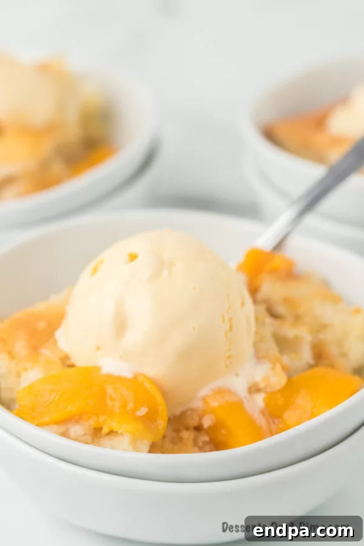 A bowl of warm peach cobbler, glistening with juicy peaches and a golden crust, generously topped with a melting scoop of vanilla ice cream.