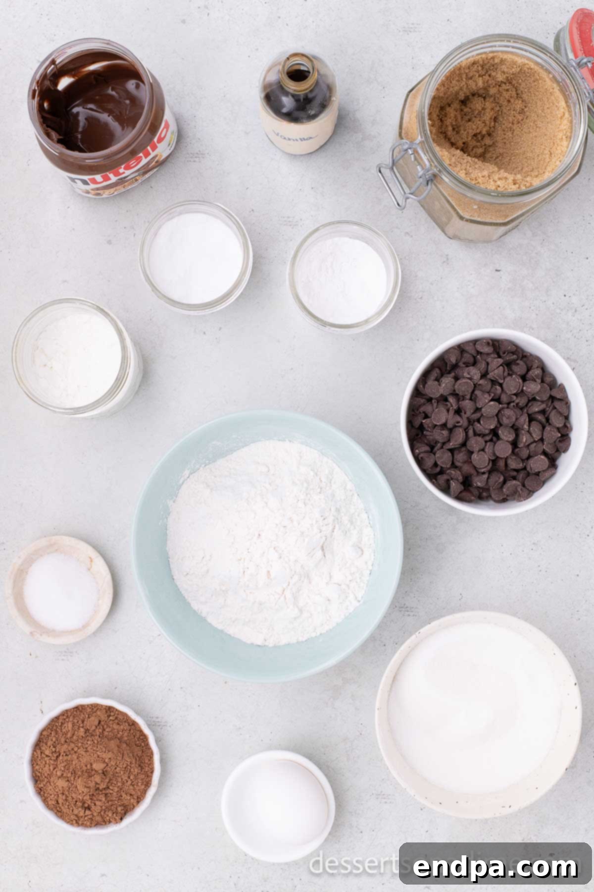 A selection of ingredients for making Nutella Cookies, including all-purpose flour, baking soda, softened unsalted butter, brown sugar, a jar of Nutella, vanilla extract, a large egg, and chocolate chips, laid out on a rustic surface.