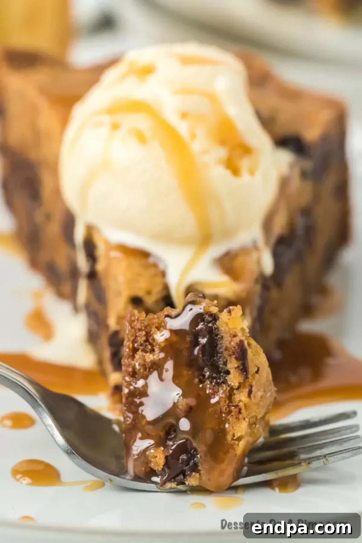 Close up image of a slice of chocolate chip cookie cake topped with ice cream on a plate, showcasing its gooey chocolate interior and golden-brown edges