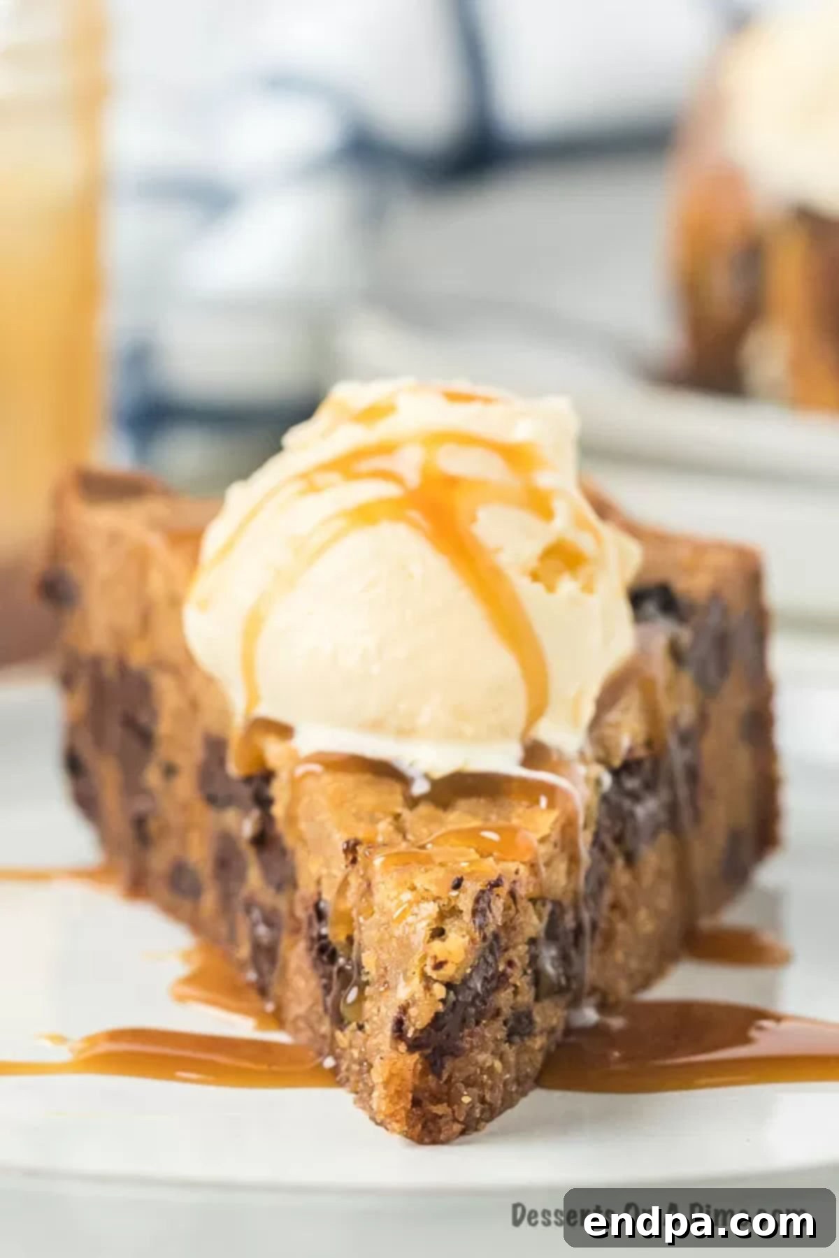 Close up image of a slice of chocolate chip cookie cake topped with ice cream on a plate, showing the rich, gooey texture and generous chocolate chips