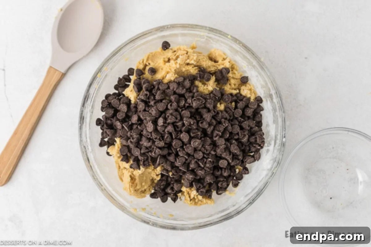 Chocolate chips being folded into the cookie dough by hand in a large mixing bowl.