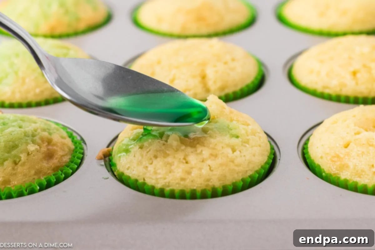 Zingy Cherry Limeade Cupcakes 6 A spoon meticulously drizzling the liquid jello mixture over the poked holes of a cupcake, with the green liquid visibly soaking into the cake.