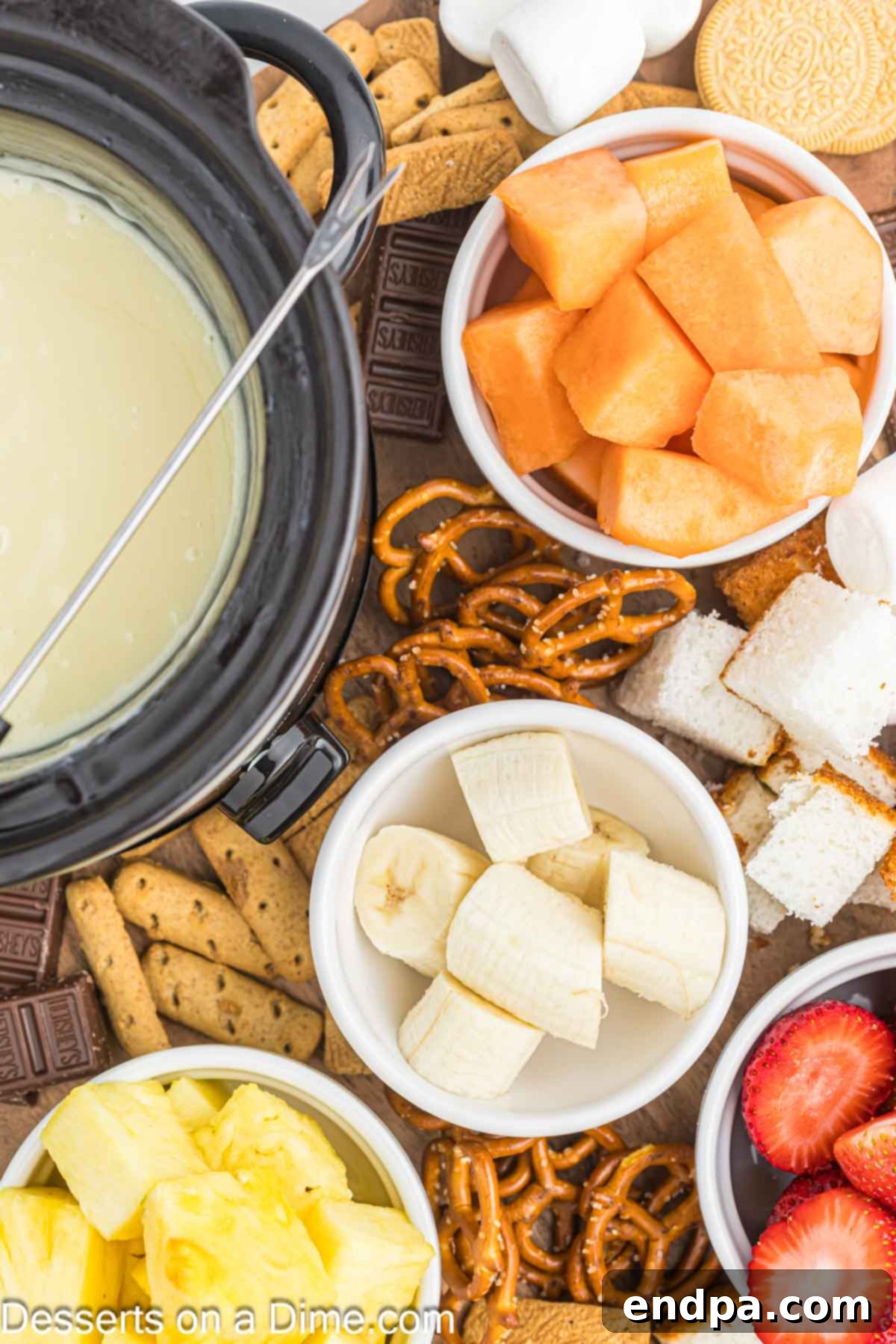 Decadent White Chocolate Fondue 7 A close-up of creamy white chocolate fondue in a pot, with a fondue fork dipping a strawberry, and various other dippers like pretzels and fruit slices in the background.
