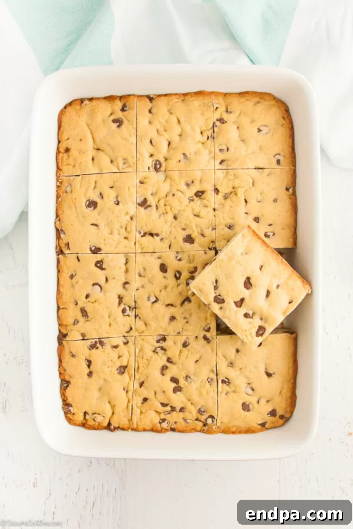 A freshly baked chocolate chip cookie cake, sliced into bars in a 9x13 inch pan. One piece is removed, showing its chewy texture and abundant chocolate chips.