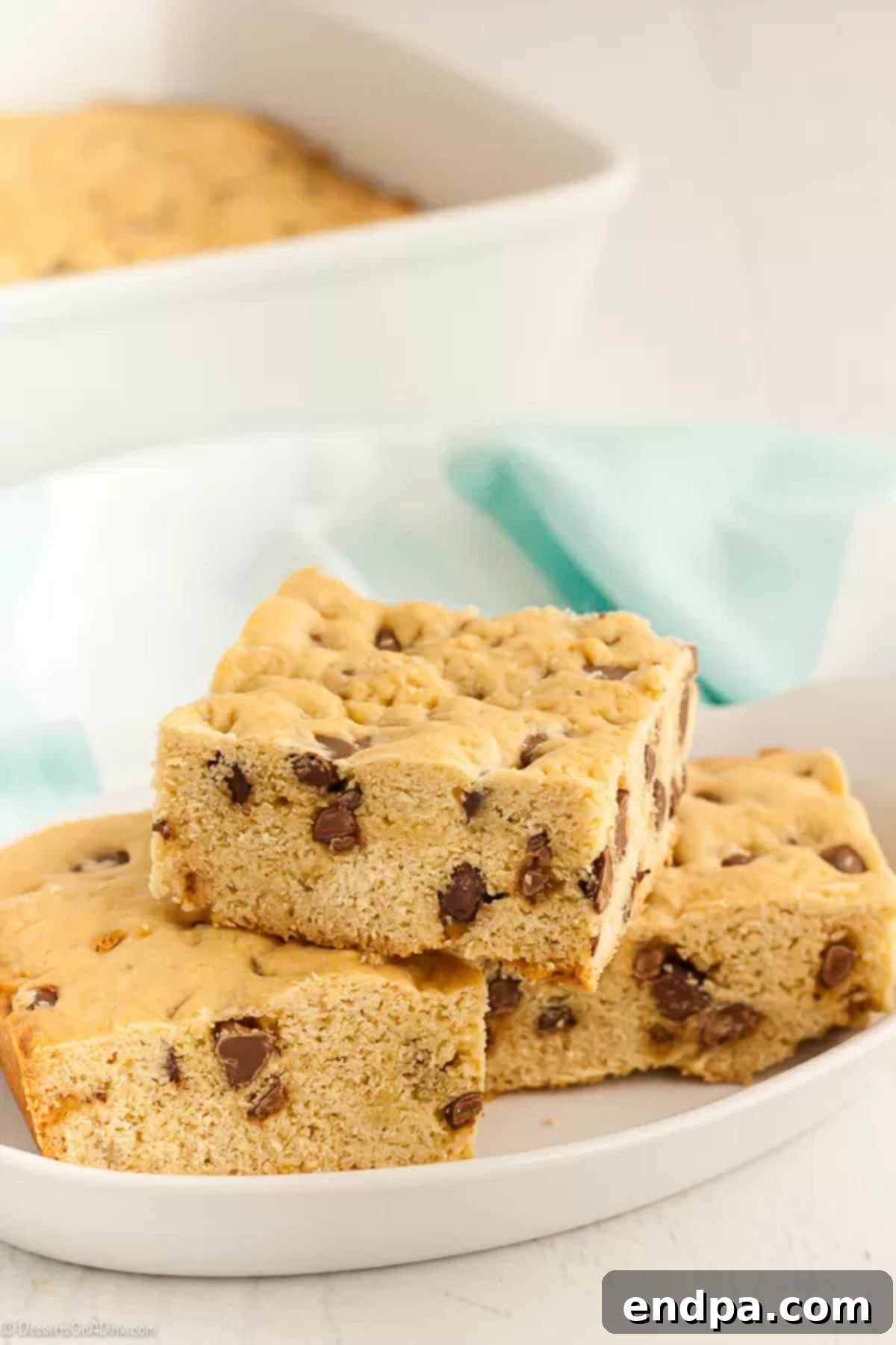 A stack of golden brown chocolate chip cookie bars on a small white platter, with the rest of the cookie cake visible in the background.