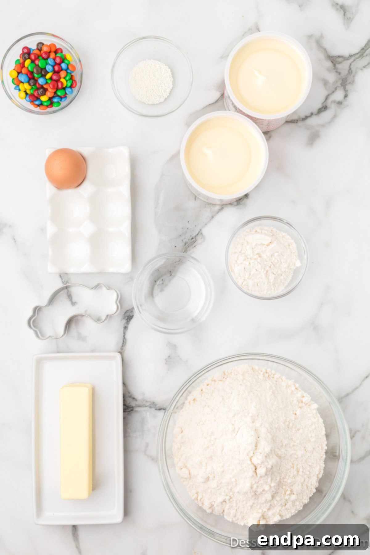 Ingredients for Rainbow Cookies laid out on a white surface: a bowl of sugar cookie mix, a stick of butter, an egg, vanilla frosting, miniature M&Ms, and white nonpareil sprinkles.